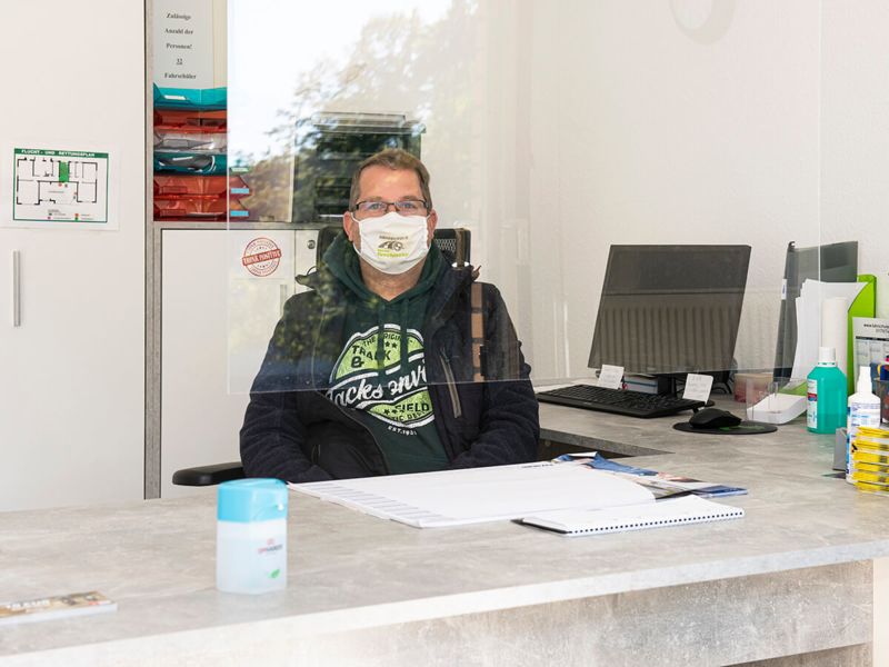 Leopoldshöher Fahrlehrer mit Mund-Nasen-Schutzmaske, hinter einer Spuckschutz Plexiglasscheibe mit Desinfektionsmitteln auf dem Schreibtisch